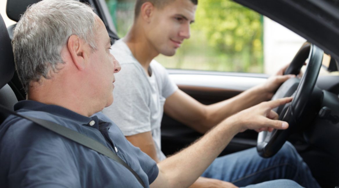 Un jeune conducteur au volant d'une voiture reçoit des instructions de son accompagnateur lors d'une session de conduite accompagnée, dans un cadre verdoyant.