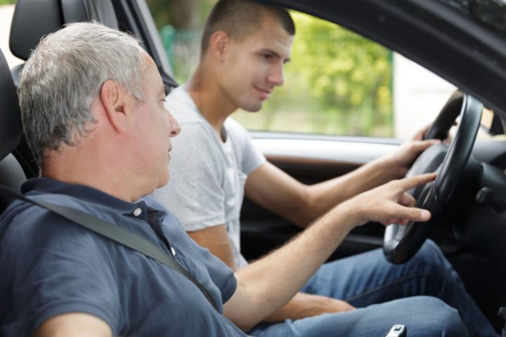 Un jeune conducteur au volant d'une voiture reçoit des instructions de son accompagnateur lors d'une session de conduite accompagnée, dans un cadre verdoyant.