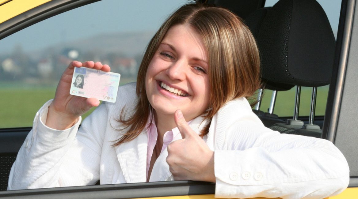 Une jeune femme souriante, assise dans une voiture jaune, montre fièrement son permis de conduire tout en levant le pouce en signe de réussite.