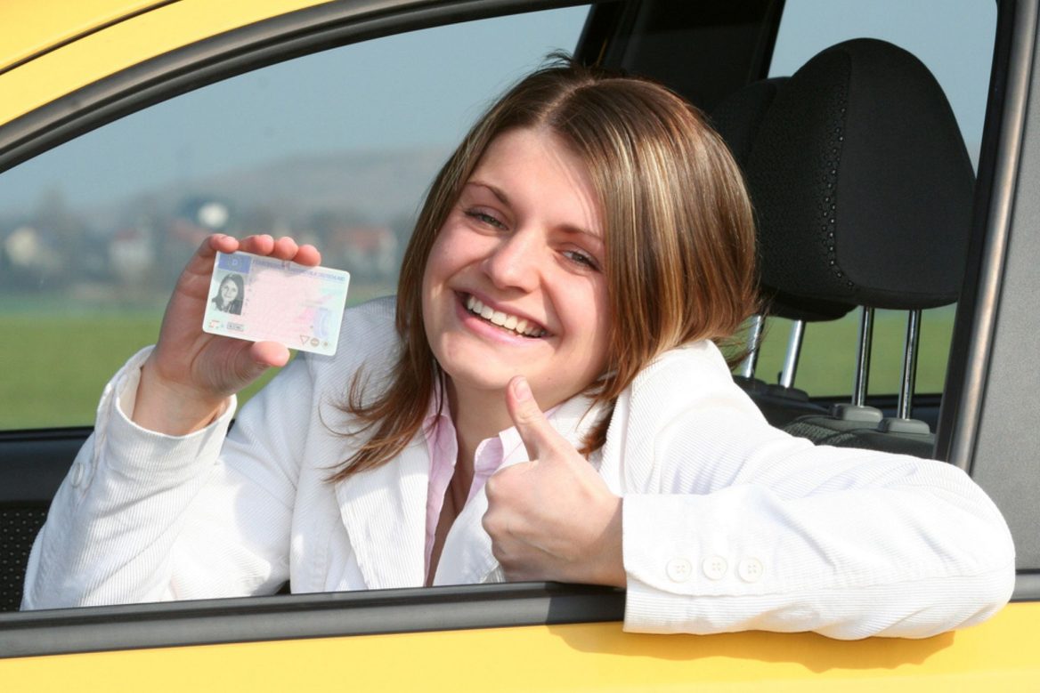 Une jeune femme souriante, assise dans une voiture jaune, montre fièrement son permis de conduire tout en levant le pouce en signe de réussite.